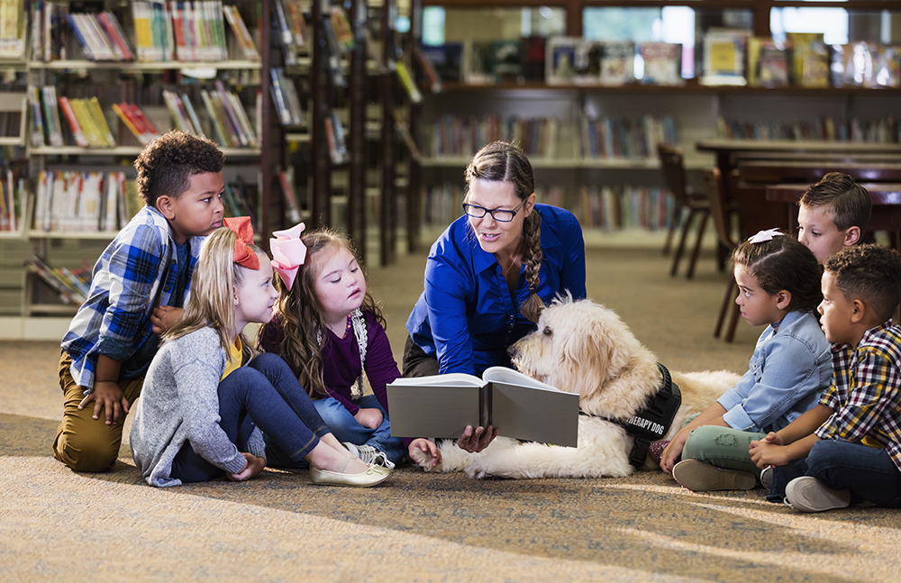 Foto: Eine Schulbegleiterin
  sitzt mit mehreren Kindern und einem Hund auf dem Boden einer Bibliothek. Sie schauen gemeinsam ein Buch an. Ein Mädchen hat das Down-Syndrom.